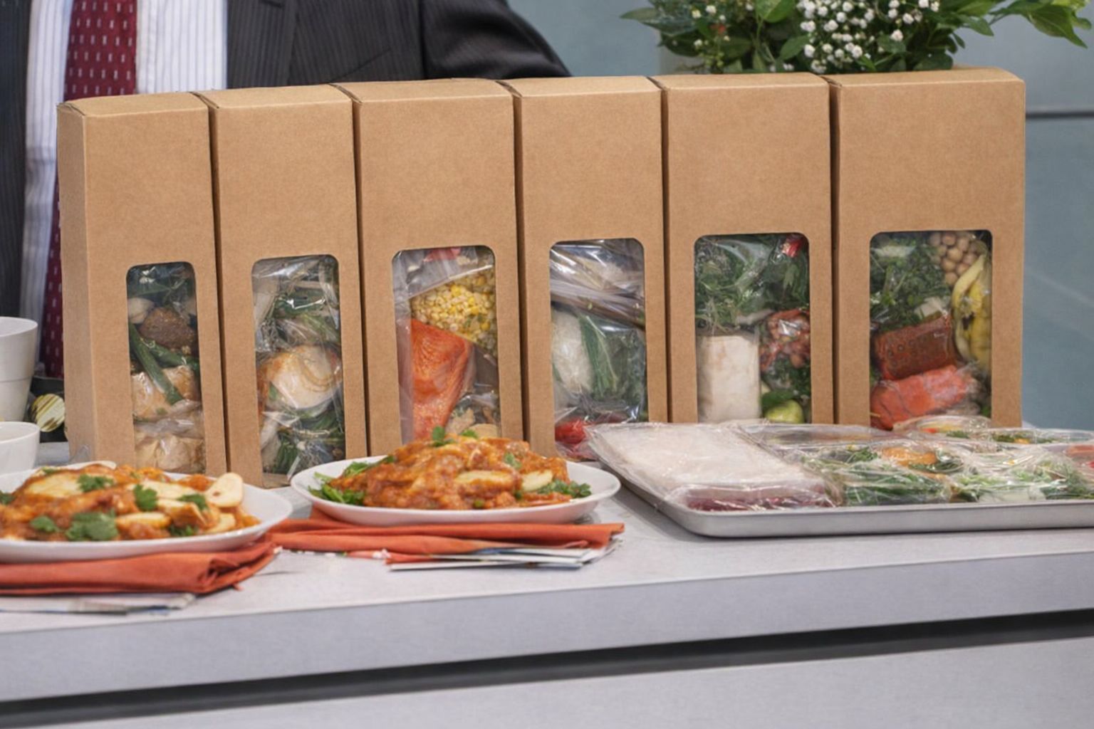 Seks måltidskasser i pap med friske råvarer og færdiglavede retter på et bord.